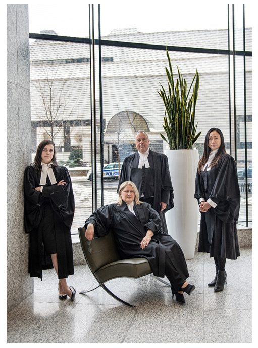The photograph is of four lawyers dressed in their formal Ontario court robes. Two women and one man are standing, and one woman is sitting on a bench in front of them. All four lawyers are posed, looking into the camera. They have serious expressions on their faces. They are in front of a large glass window and a large decorative white vase with a green tropical plant. In the background, outside, is the front of the Ottawa Courthouse and an Ottawa Police Services cruiser.